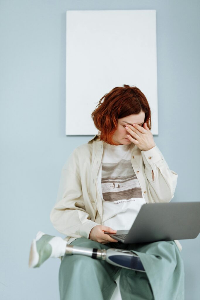 Woman with prosthetic leg sits indoors using a laptop, expressing frustration.
